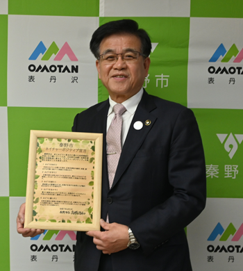 秦野市のバックパネルの前で、額に入った「秦野市ネイチャーポジティブ宣言」を両手で持っている市長の写真