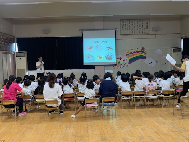 白いTシャツ姿の講師が室内で、椅子に座った大勢の園児たちと数名の大人を前に、スクリーンに水辺の生き物のイラストと「みずなしがわ」というタイトルが投影されたエコスクールの授業の様子の写真