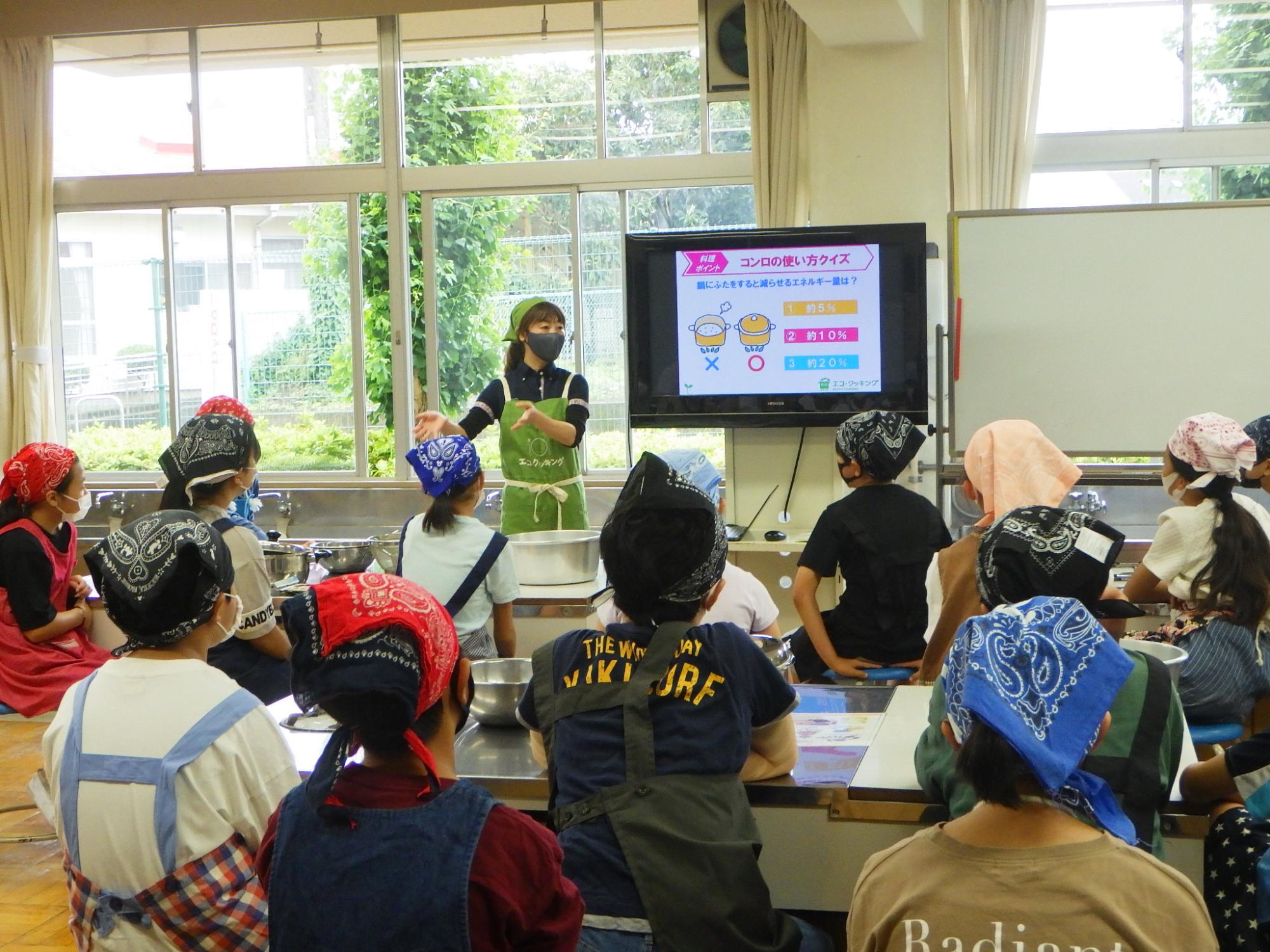 大きなモニターが設置された大きな窓のある調理室のような場所で、エプロンとバンダナを着用した多くの児童たちが、調理実習の指導者と思われる女性がコンロの使い方に関する画面を見ながら座っている授業の様子の写真