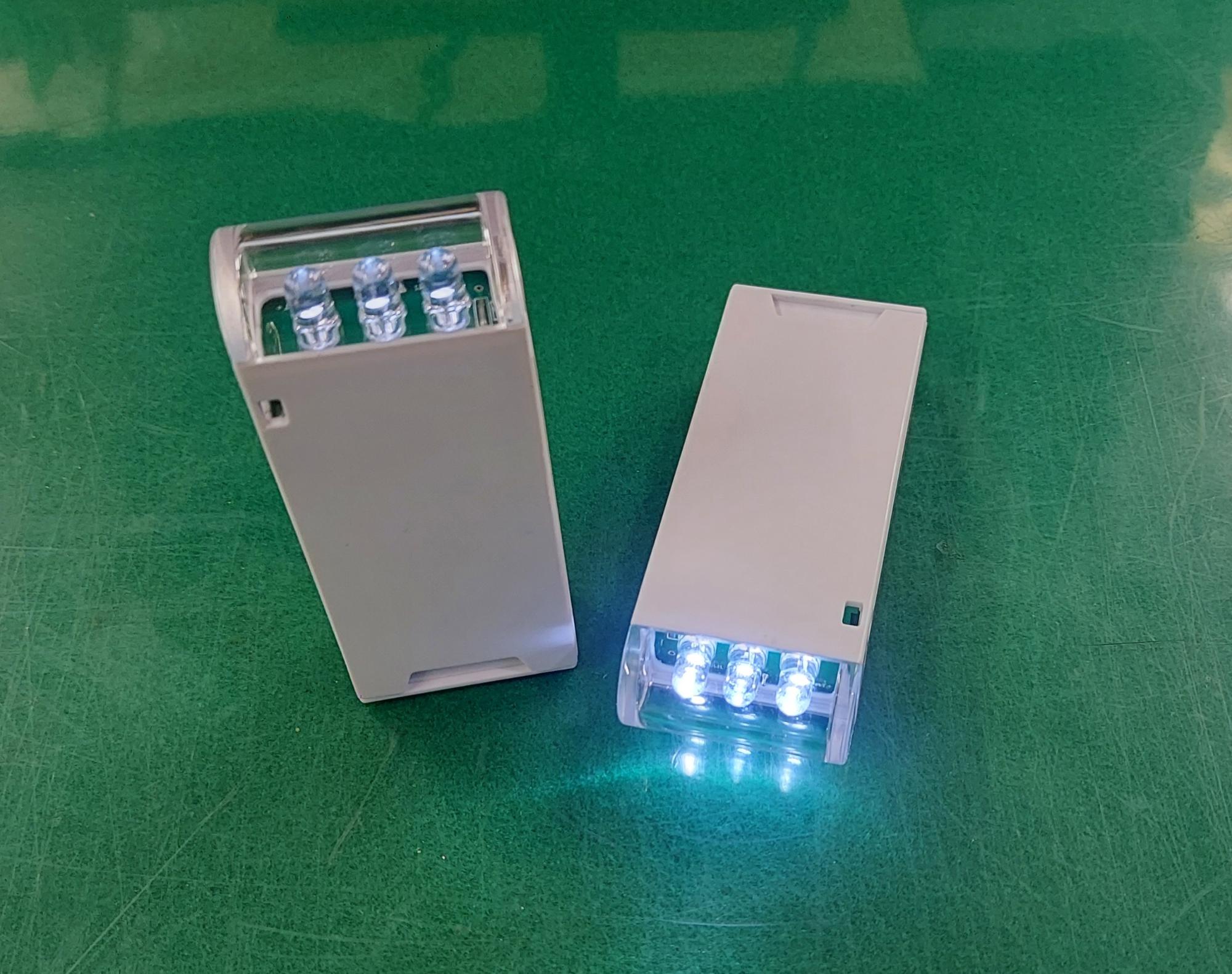 緑色の作業台のような場所に置かれた、白色の長方形のプラスチック製筐体を持つ2つの小型ライトの写真で、左側のライトは消灯し、右側のライトは上部の透明カバーから青白い3つのLEDが強く発光している写真