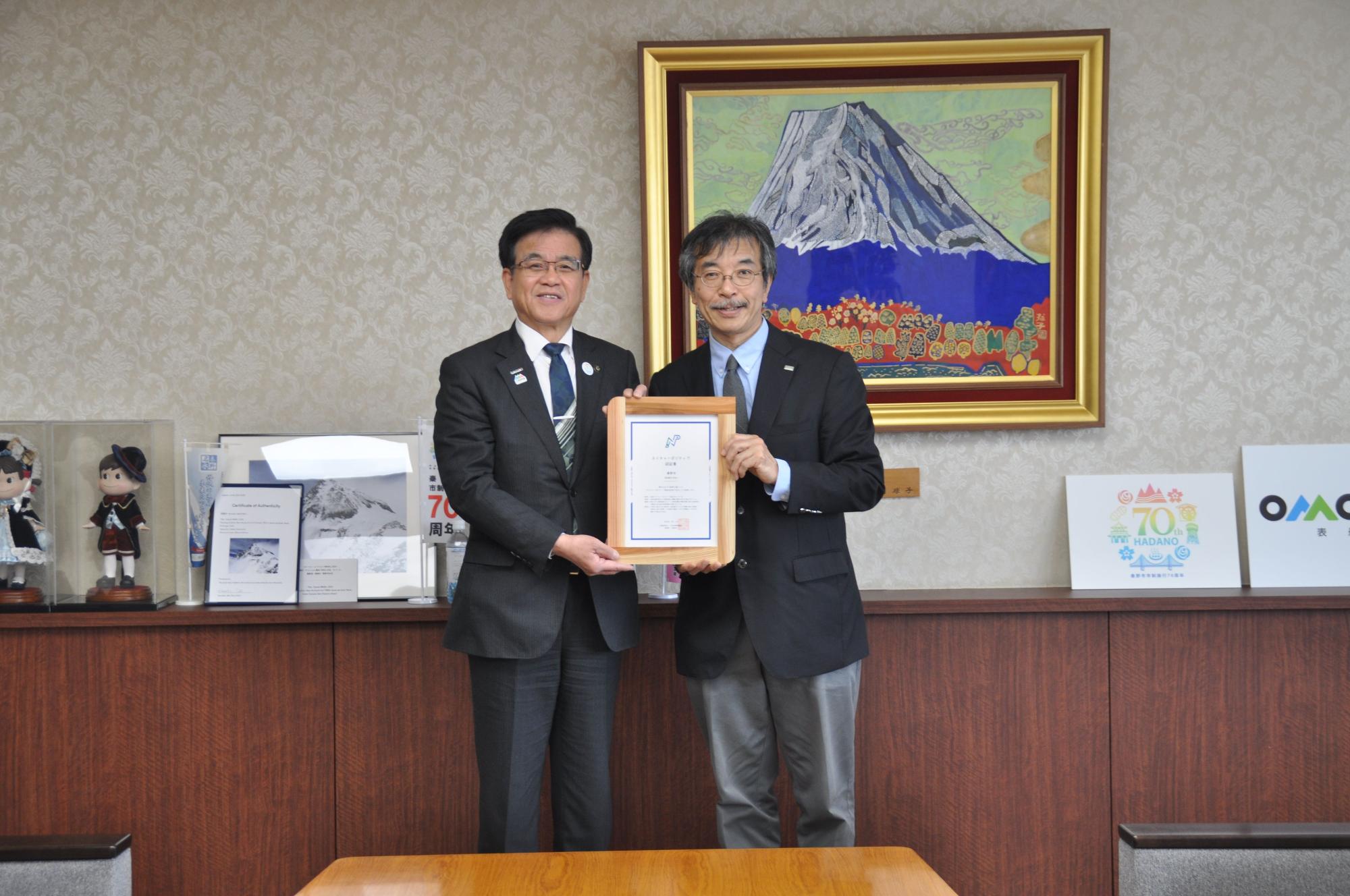 金色の額縁に入った山頂に雪が積もった大きな山の絵画が壁に飾られた落ち着いた背景の部屋で、土屋理事長と市長が額に入ったネイチャーポジティブ認証書を一緒に持ち並んでいる記念写真
