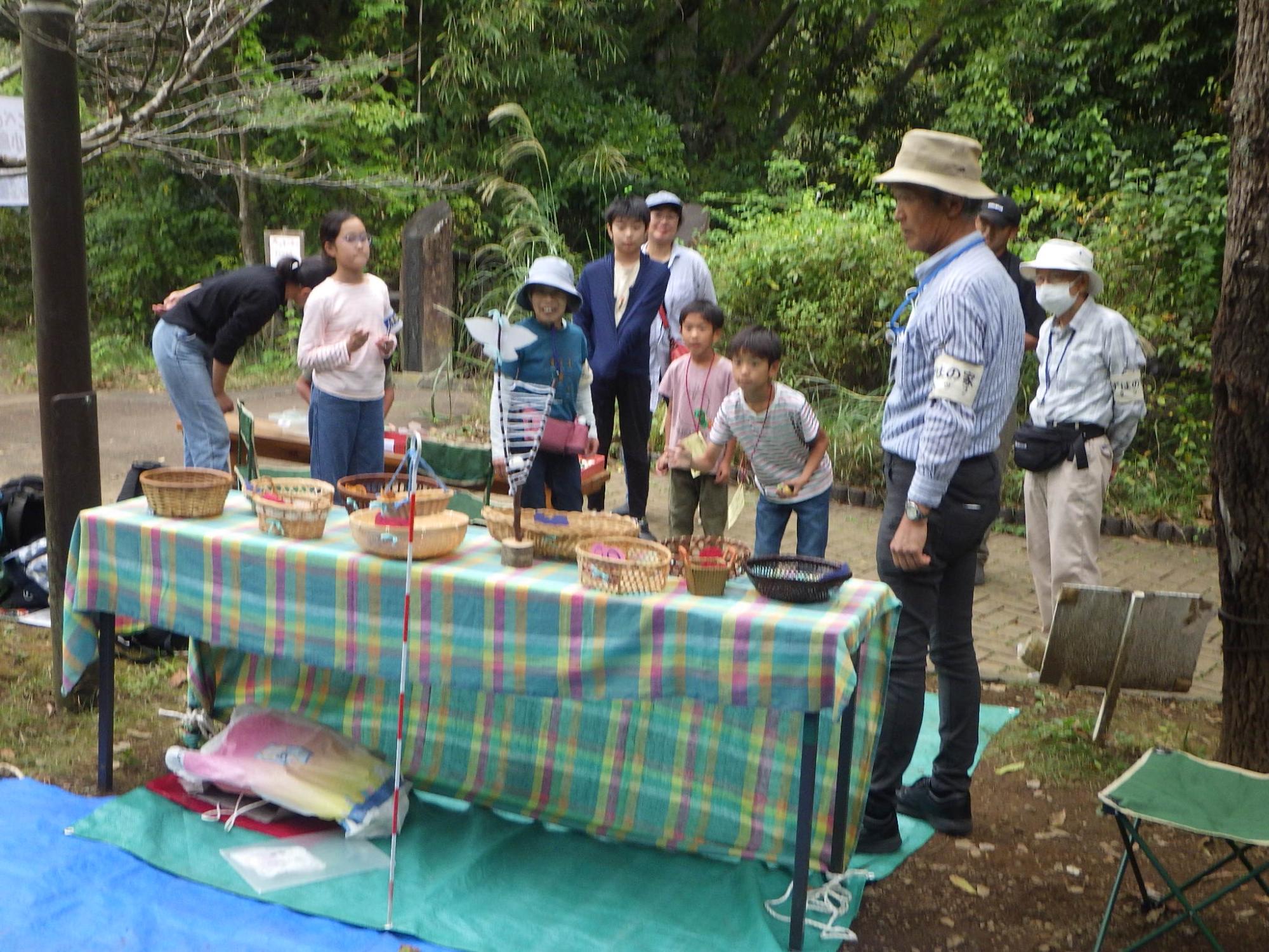 公園内の自然の中、チェック柄のテーブルクロスが敷かれたテーブルを囲み、帽子をかぶったスタッフと数人の子供たちが、竹とんぼやこまと思われる手作りの遊び道具で楽しそうに遊んでいる様子の写真