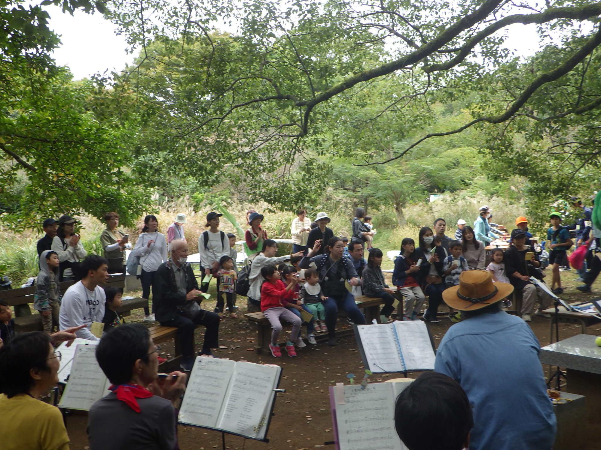豊かな緑に囲まれた屋外で、楽譜立ての奥に座る演奏者が管楽器を演奏している音楽を多くの参加者が木のベンチに座って観覧している写真