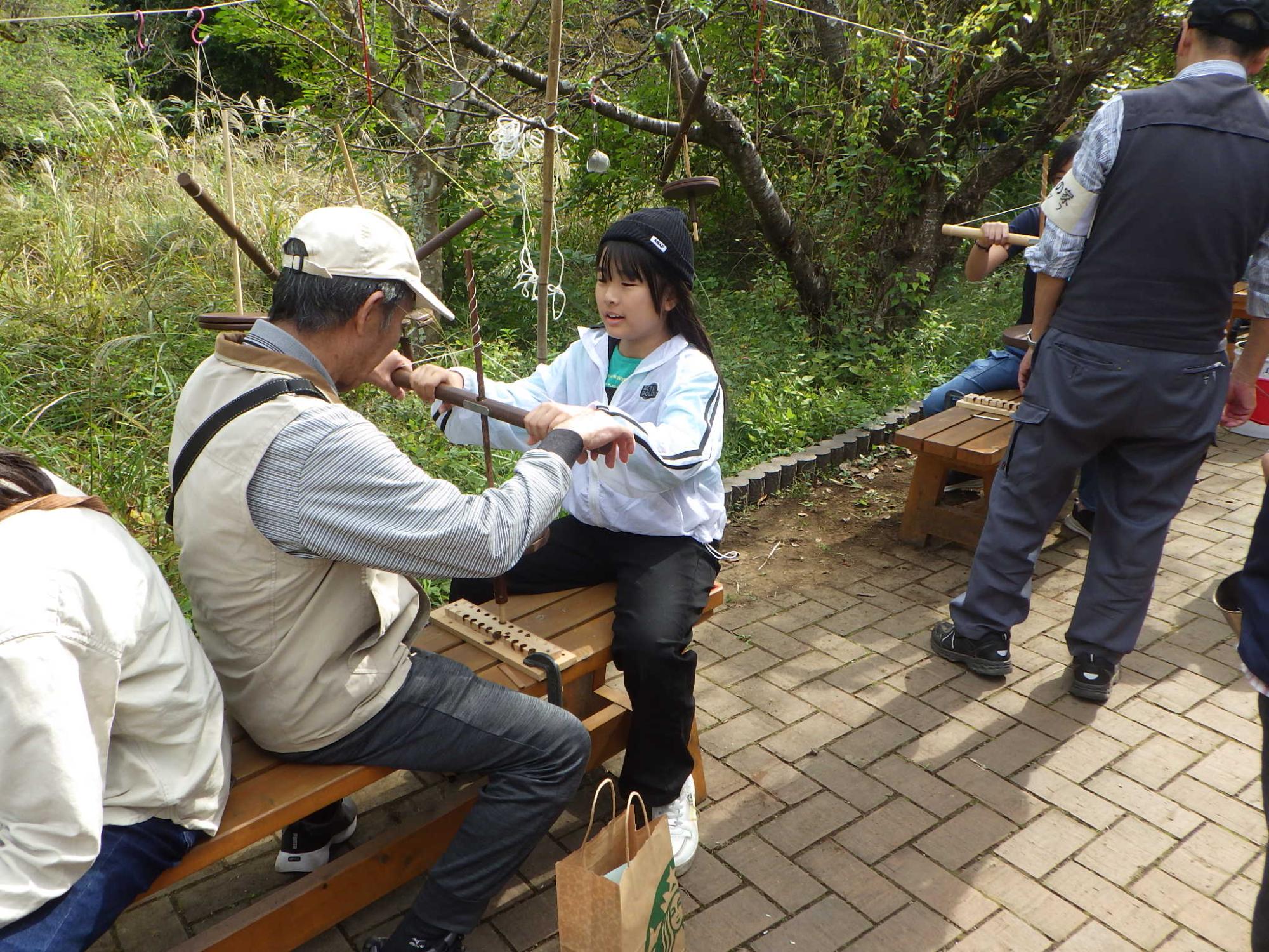 屋外でベンチに座った大人の男性が、火おこし器のような道具の棒を両手で持って座っている女の子に正しい使い方を指導している様子の写真
