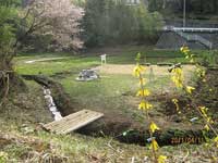 奥には白いガードレールや階段が見え、手前の土手の斜面に小さな水路が流れ、細い木の板が橋のように架けられている自然豊かな風景写真