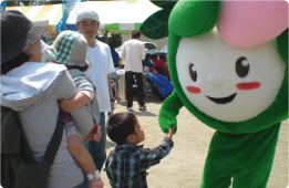 屋外イベント会場で、母親に抱かれた幼児と、チェック柄の服を着た別の子供が、緑色とピンク色の大きな花のような顔を持つかなりんちゃんと握手している様子の写真