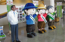 屋内会場で、白い上着を着た男性が左端に立ち、その右側には「全国植樹祭」と書かれたタスキをかけた丹沢はだの三兄弟が並んで手を挙げている、イベントの一場面の写真