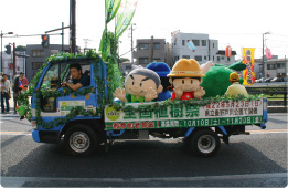 緑の葉で飾られた青いトラックの荷台に、「全国植樹祭」の文字と日程が書かれた幕とともに、丹沢はだの三兄弟とかなりんちゃんが乗って手を振っているパレードの写真