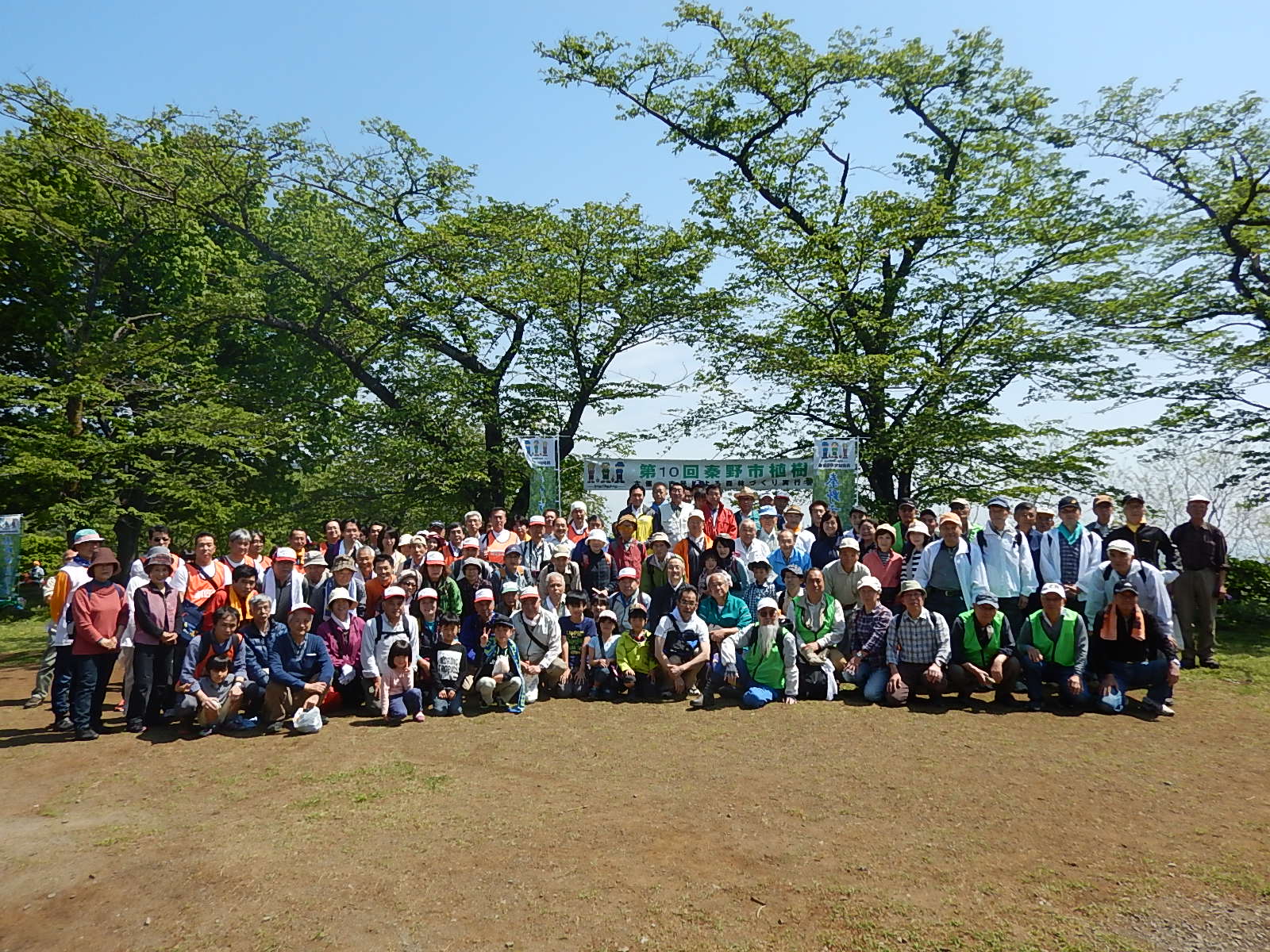 澄み切った青空の下、広場の中央で「第10回秦野市植樹祭」と書かれた横断幕を背に、参加者たちが集まって並び、笑顔でポーズをとっている植樹祭の記念集合写真