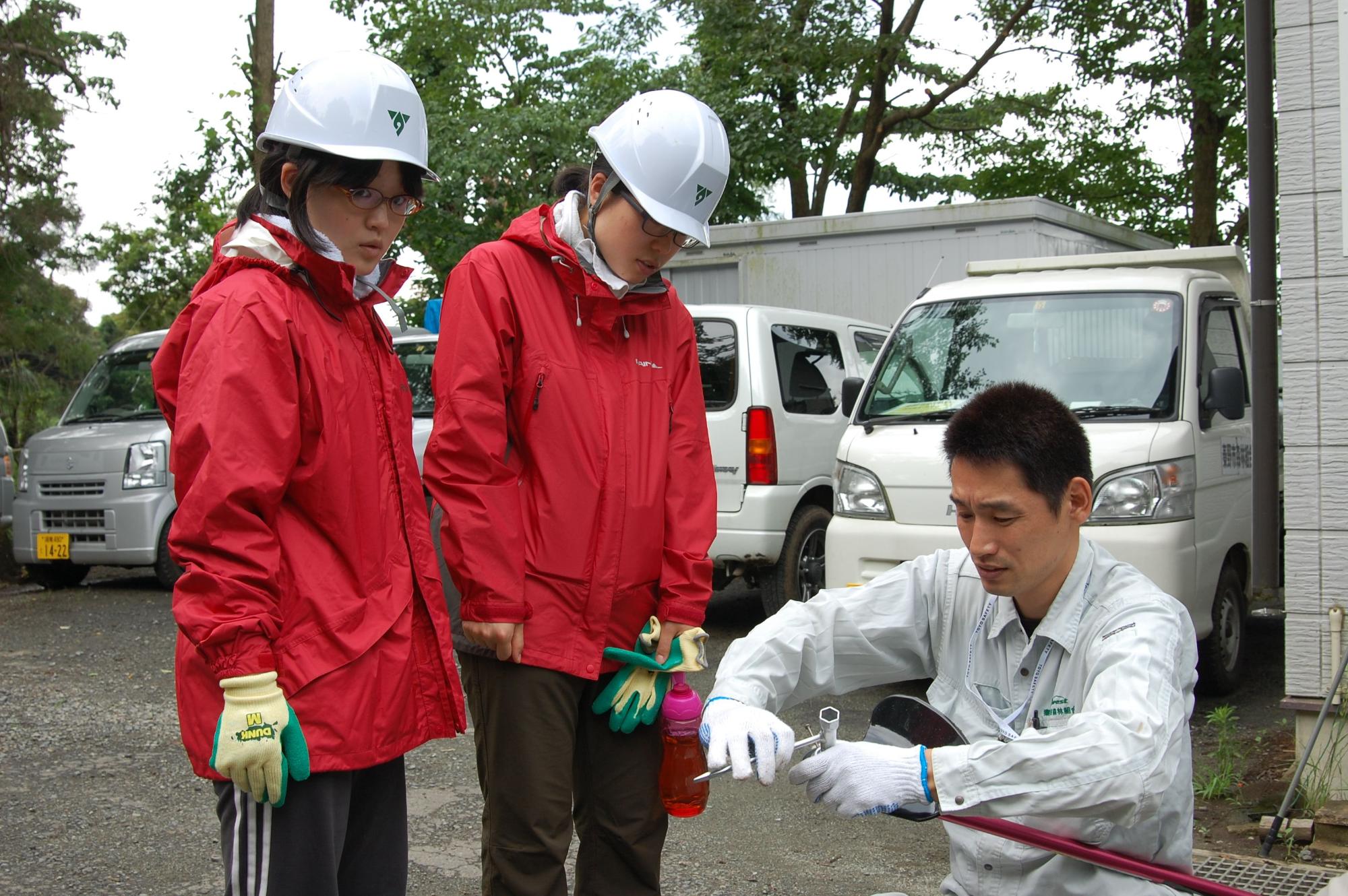 白いヘルメットと赤い雨具を着用した2人の女性が立って見守る中、作業服姿の男性が工具を使ってしゃがみこみパイプのようなものを組み立てている屋外での研修風景の写真