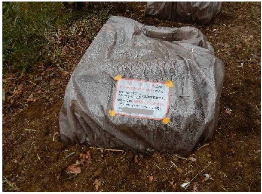 グレーのシートに包まれ地面に置かれた大きな袋のような塊に、赤と黒の文字で書かれた「注意」の紙が貼り付けられており、土壌の上でくん蒸処理がされている現場の写真