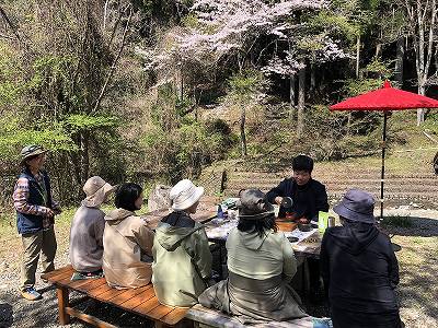 春の木漏れ日が差す屋外で、ベンチに座ったハイキング姿の6人が、中央でコーヒーを淹れている男性を囲み、手前には赤い野点傘が見える中で休憩しているイベントスナップ写真