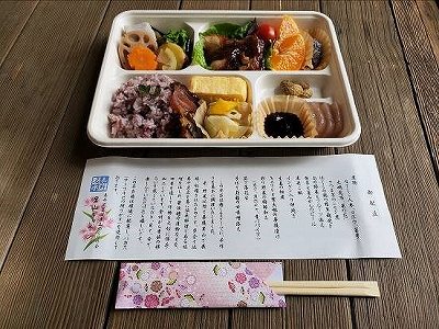 木目調のテーブルの上に置かれた、複数の仕切りに紫色の混ぜご飯、玉子焼き、煮物、黒豆などが入った色鮮やかな和風の弁当と、料理の説明書き、桜柄の箸袋に入った割り箸が並ぶ料理写真