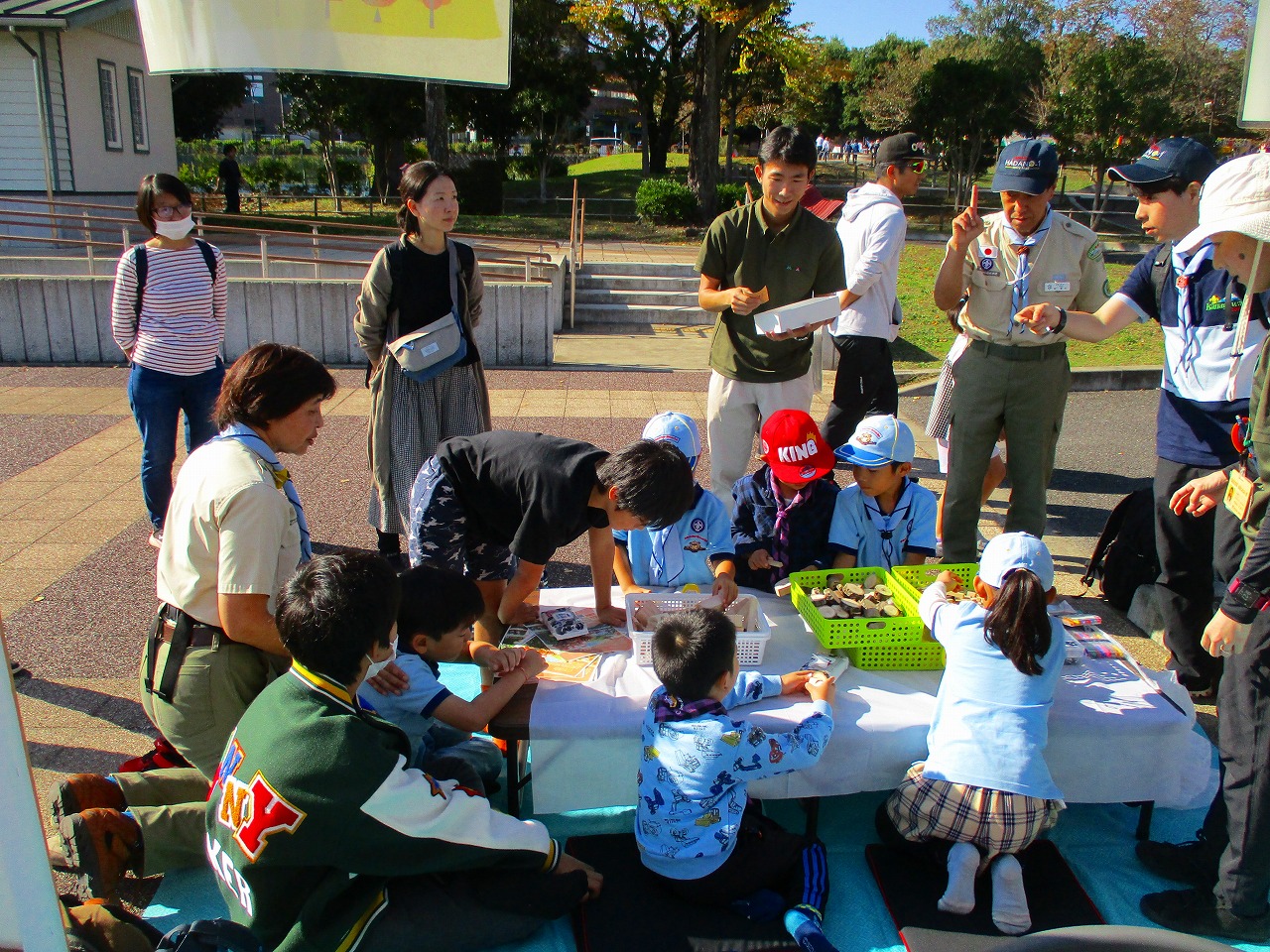晴れた屋外の広場で、カブスカウトの制服を着た子供たちがテーブルを囲んで何かを学んだり作業したりしており、周りを保護者らしき大人や指導者が立ったりしゃがんだりしながら見守るイベントの様子の写真