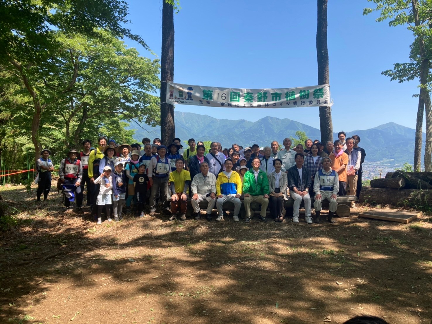 抜けるような青空と遠くの山々を背景に、第16回秦野市植樹祭の横断幕を掲げた木々の下で、多数の参加者が集まり、笑顔でカメラに向かってポーズをとっている植樹祭の集合写真