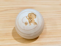 木目調の明るい木製土台の上に置かれた、表面に木製玩具のキャラクターと見られる楽しそうな表情の木製のきのこのイラストがレーザー刻印されている、表面が滑らかな丸いヒノキ玉の写真