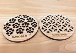 「OMOTAN」という文字がレーザー刻印されている、右側が幾何学的な星やダイヤの模様、左側が花柄の透かし彫りされた2枚の薄い木製の円形コースターが並んでいる工芸品の写真