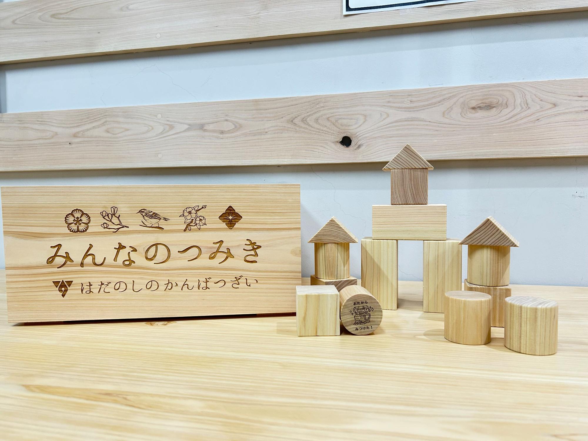 「みんなのつみき」と刻印された木製の看板と、三角屋根や円柱などの様々な形のシンプルな木製積み木が、木目のある床と木製パネルの壁の前に並べられている、温かみのある木製玩具の写真