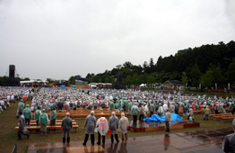 雨が降りしきる曇天の野外会場で、緑のTシャツやカッパを着た大勢の参加者がベンチに座ったり立ち見したりしているイベント会場の写真