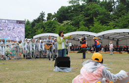 野外ステージ前の芝生で、マイクを持った女性が歌い、その背後にはギターを持った男性やカッパ姿の参加者が並び、左側の大型ビジョンには群衆の様子が映し出されている写真