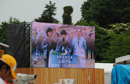 野外イベント会場に設置された大型ビジョンに、男女3人が映し出されている写真