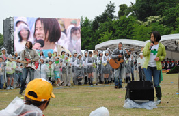 野外イベントの芝生の上で、エレキギターとアコースティックギターを抱えた男性たちの演奏に合わせて女性が歌っており、後方にはカッパ姿の大勢の参加者がその歌を聞いている様子の写真