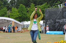 野外の芝生の上で、薄い黄緑色のジャンパーを着てマイクを握った女性が両手を高く上げている写真