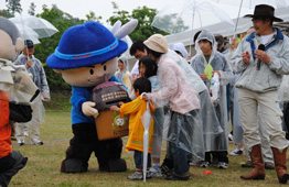 雨の野外イベントで、青い帽子をかぶったマスコットキャラクターが募金箱を持って立ち、黄色いカッパ姿の小さな子供が母親と一緒に寄付をしている瞬間を捉えた写真