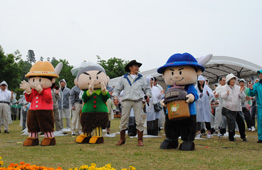 曇り空の野外イベントで、赤い服、緑の服、青い服の3体の着ぐるみキャラクターが並び、中央にはカウボーイハットを被った男性が立ち、後方では多くの参加者が拍手をしている写真
