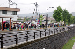 黒い手すりの石積み護岸沿いの歩道に、傘をさした大勢の人々が長く一列に並んで立っており、天皇皇后両陛下が車で通るのを待っている様子の写真