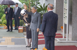 雨の屋外で、天皇陛下が片手を上げて微笑みながら挨拶をされ、隣の天皇皇后は頭を下げられ、近くには天皇皇后両陛下を出迎えるスーツ姿の男性が立っている写真