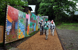 雨が降る屋外で、カラフルな絵画が描かれた壁が道沿いに並んでおり、その道をカッパを着用した人々が歩いている写真