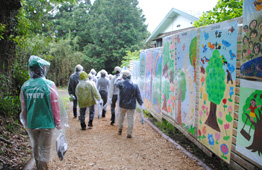 幼稚園・小学校・中学校の生徒が描いたメッセージボードが並ぶ壁の横の道を人々が歩いている写真