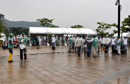 雨が降る広場で、緑色のベストや白いカッパを着た多くの人々が、大きな白いテントの周りに集っている様子を捉えた写真