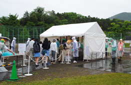 雨の屋外イベント会場で、白いテントの前に大勢の参加者やスタッフが並び、入場ゲートで列を作っている写真