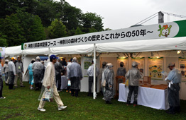 雨の降る屋外イベント会場で、「神保森林認証ブース ～神奈川の森林づくりの歴史とこれからの50年～」という看板を掲げた白いテントに、カッパを着た多くの人々が集まっている写真