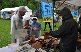 雨が降る屋外イベントのテントの下で、カッパを着た大人や子供が、木片や工作物が並べられたブースを興味深そうに見ている写真