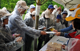 雨の屋外イベントで、カッパを着た参加者がテントの下で配布物を受け取り、周辺が賑わっている写真