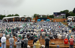 雨の降る野外会場で、透明なカッパを着用した非常に大勢の参加者が木製ベンチに座ってステージを見ており、右側の大型ビジョンには映像が映し出されている写真