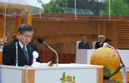 木製の壁を背景にした式典会場で、衆議院議長・大会会長が白い演台で原稿を読み上げており、演台の横には巨大な地球儀のようなオブジェが置かれ、後方には天皇皇后両陛下が座っている写真