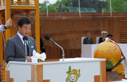 木製の壁を背景にした式典会場で、神奈川県知事が白い演台で原稿を読み上げており、演台の横には巨大な地球儀のようなオブジェが置かれ、後方には天皇皇后両陛下が座っている写真