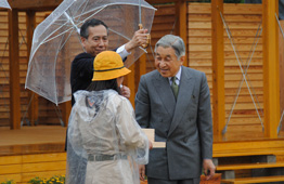 雨の中、透明のカッパと黄色の帽子を着用した若い女性が木の箱のようなものを手に持っており、向かい側に立たれた天皇陛下が笑顔で微笑んでいる写真