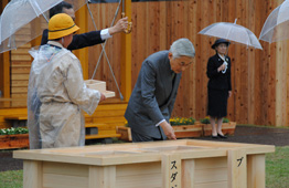 雨の降る屋外式典で、天皇陛下が、正面にある大きな木箱の台に身をかがめてお手播きされている写真