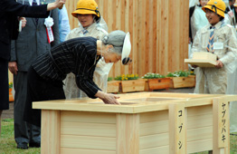 雨の降る屋外式典で、天皇皇后が、正面にある大きな木箱の台に身をかがめてお手播きされている写真