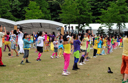 曇り空の野外イベント会場の芝生の上で、カラフルなTシャツやスパッツなどの派手な衣装を着た大勢の子供たちが、並んで両手を上げてダンスをしている写真