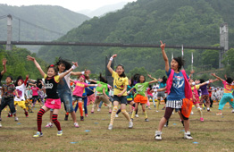 広い芝生の会場で、カラフルで個性的な衣装を身にまとった大勢の子供たちが、一斉に両手を広げて踊っている写真