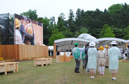 雨の野外式典会場の芝生で、透明や緑色のカッパを着た人々が立ち並び、左側の大型ビジョンには地球儀のオブジェに触れる様子が映し出されている写真