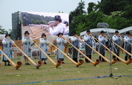 曇天の野外イベントの芝生の上で、伝統衣装らしき服装を着た男女が、長く湾曲した木製のアルプホルンを地面に立てて一列に並んで演奏し、後方の大型ビジョンにも演奏者の姿が映し出されている写真
