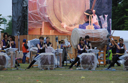 曇天の野外会場で、紫色の衣装を着た和太鼓演奏グループが、ビニールシートで覆われた大太鼓や中太鼓を打ち鳴らし、後方の大型ビジョンにも演奏の様子が映し出されている写真