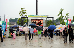 雨が降る屋外イベント会場で、多くの参加者が傘をさして中央の大型ビジョンを囲むように立っている写真